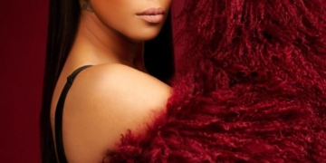 R&B singer Monica holding up a plush red coat to the side of her face, posing against a red background. She is wearing a matching red headband and elaborate eye makeup, looking directly at the camera.