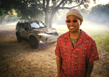 Anderson Paak wearing a patterned red shirt and sunglasses, paired with a beige hat, is smiling while standing outdoors. Behind him, a 2024 Lexus GX is parked amid a backdrop of trees illuminated by soft sunlight, with a mist of dust in the air
