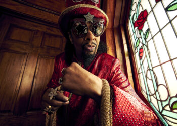 A photo of Bootsy Collins wearing hat and sunglasses poses in front of colorful stained glass window.