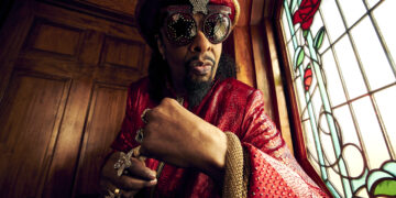 A photo of Bootsy Collins wearing hat and sunglasses poses in front of colorful stained glass window.