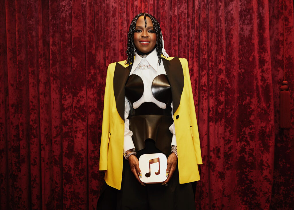 Lauryn Hill stands against a red curtain backdrop holding a white award with a red music note symbol on it. She wears a stylish outfit featuring a black and white corset top under a yellow and black jacket, complemented by her braided hair adorned with beads.