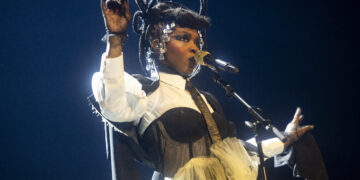 Lauryn Hill in an elaborate costume singing into a microphone on a concert stage, highlighted by dramatic stage lighting. The costume features a distinctive headdress and structured outfit with Victorian influences.