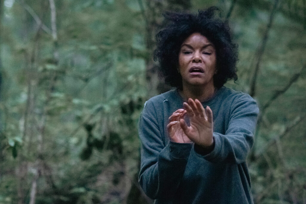 LeToya Luckett in Lifetime movie I Thought My Husband's Wife Was Dead, pictured with a fearful expression running through a wooded area, hands up defensively. Their dark hair is disheveled, and they are wearing a gray long-sleeve shirt. The backdrop is a densely wooded forest, creating a moody atmosphere.