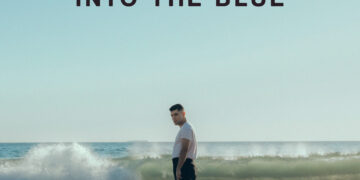 Album cover for Aaron Frazer's "Into the Blue," featuring the artist standing on a beach with waves in the background under a clear sky.