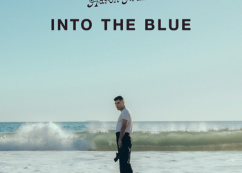 Album cover for Aaron Frazer's "Into the Blue," featuring the artist standing on a beach with waves in the background under a clear sky.