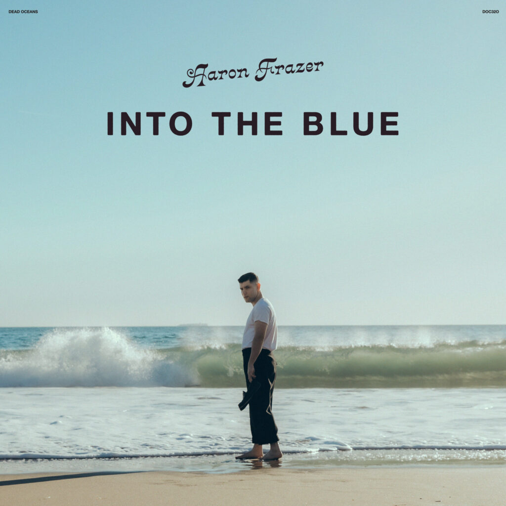 Album cover for Aaron Frazer's "Into the Blue," featuring the artist standing on a beach with waves in the background under a clear sky.