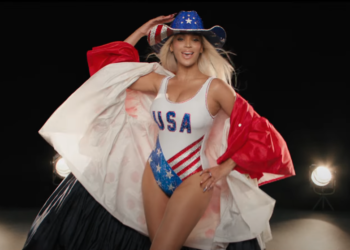 Beyoncé wearing a patriotic outfit themed with the USA flag colors, including a blue cowboy hat with stars, a one-piece swimsuit, and a flowing cape. She is are posing confidently in front of a dark background lit by studio lights. The swimsuit is emblazoned with "USA" across the chest.