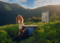 A cover art for BLK ODYSSY's album titled "1-800-FANTASY" featuring two individuals in a surreal outdoor setting. The scene includes lush green hills under a vibrant sky. One person wearing a white dress is standing and looking off into the distance, while the other, wearing a suit, sits on the ground with their head resting on a sideways old-fashioned tube television. Near them is a classic phone booth. The image includes the advisory notice "PARENTAL ADVISORY EXPLICIT CONTENT".