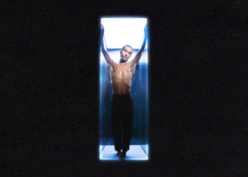 Forest Claudette poses shirtless in a blue-lit, vertical capsule, arms raised and eyes closed, set against a dark background.