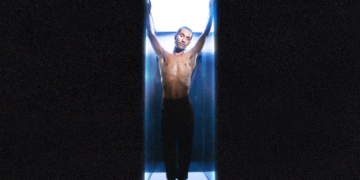 Forest Claudette poses shirtless in a blue-lit, vertical capsule, arms raised and eyes closed, set against a dark background.