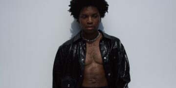 R&B singer Gallant with short curly hair stands against a plain light-colored wall. He is wearing a black leather jacket over an unbuttoned shirt, revealing a bare chest. A necklace with the word "ZINC" is visible around his neck.