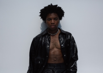 R&B singer Gallant with short curly hair stands against a plain light-colored wall. He is wearing a black leather jacket over an unbuttoned shirt, revealing a bare chest. A necklace with the word "ZINC" is visible around his neck.