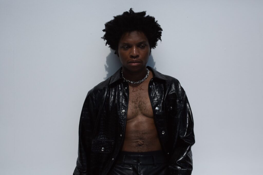 R&B singer Gallant with short curly hair stands against a plain light-colored wall. He is wearing a black leather jacket over an unbuttoned shirt, revealing a bare chest. A necklace with the word "ZINC" is visible around his neck.