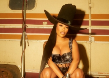 K. Michelle, who is gearing up to release her country album, in a cowboy hat and plaid crop top squatting in front of an old camper trailer at night.
