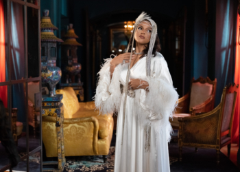 Marsha Ambrosius dressed in an elegant white outfit adorned with feathers and beads stands in a luxuriously decorated room, featuring rich blue walls and eclectic furnishings. Marsah Ambrosius is accessorized with a feathered headpiece and holds a thin, beaded accessory to their face. The ambient lighting gives the setting a warm, opulent atmosphere.