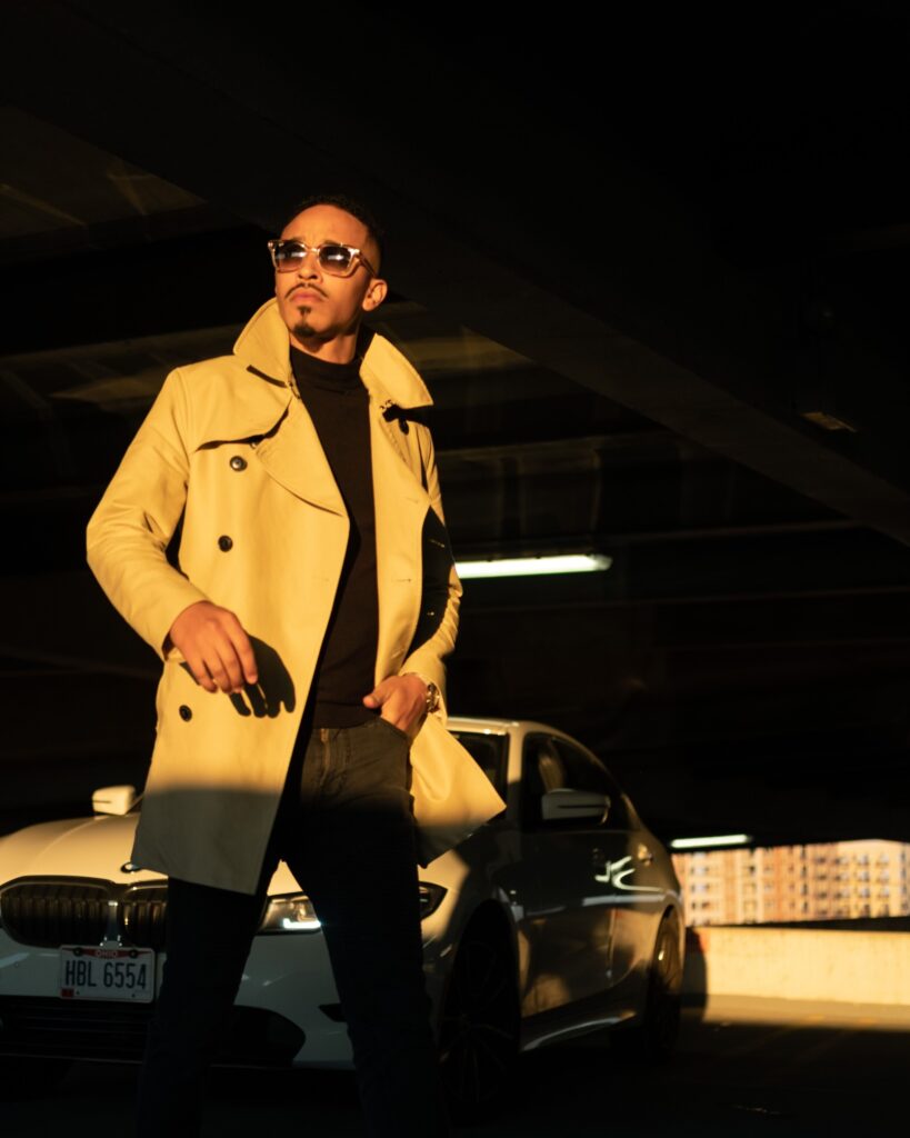 A photo of R&B singer October London in a beige trench coat and sunglasses stands confidently in a parking garage, next to a BMW car, under dramatic yellow lighting.