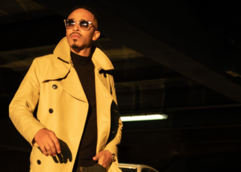 A photo of R&B singer October London in a beige trench coat and sunglasses stands confidently in a parking garage, next to a BMW car, under dramatic yellow lighting.