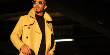 A photo of R&B singer October London in a beige trench coat and sunglasses stands confidently in a parking garage, next to a BMW car, under dramatic yellow lighting.