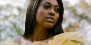 Close-up portrait of Jazmine Sullivan wearing a yellow tulle outfit, gazing off to the side with a serene expression, framed against a softly blurred natural background.