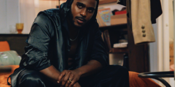 A photo of R&B singer TA Thomas wearing a black outfit, sitting on an orange couch in a warmly lit room.