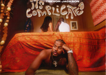 This is an album cover for "It's Complicated" by Trevor Jackson. The image depicts Trevor Jackson lying on a red bed, gazing directly at the camera with an intense expression. In the background, a couple is visible under a sheer red fabric, which adds a layer of complexity to the scene. Above the bed hang wooden beads, casting patterns on Jackson and the drapery. The cover contains explicit content warning and artist's name in bold letters.