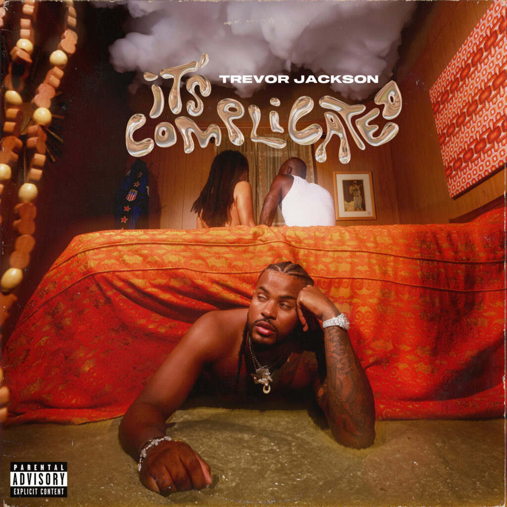 This is an album cover for "It's Complicated" by Trevor Jackson. The image depicts Trevor Jackson lying on a red bed, gazing directly at the camera with an intense expression. In the background, a couple is visible under a sheer red fabric, which adds a layer of complexity to the scene. Above the bed hang wooden beads, casting patterns on Jackson and the drapery. The cover contains explicit content warning and artist's name in bold letters.