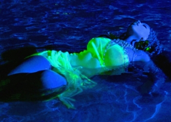 The cover art for Chloe Bailey's album Trouble in Paradise. She is floating in water, illuminated by blue and green lights, wearing a glow-in-the-dark costume resembling a mermaid tail and bikini top. The reflective details on the costume sparkle subtly under the light.