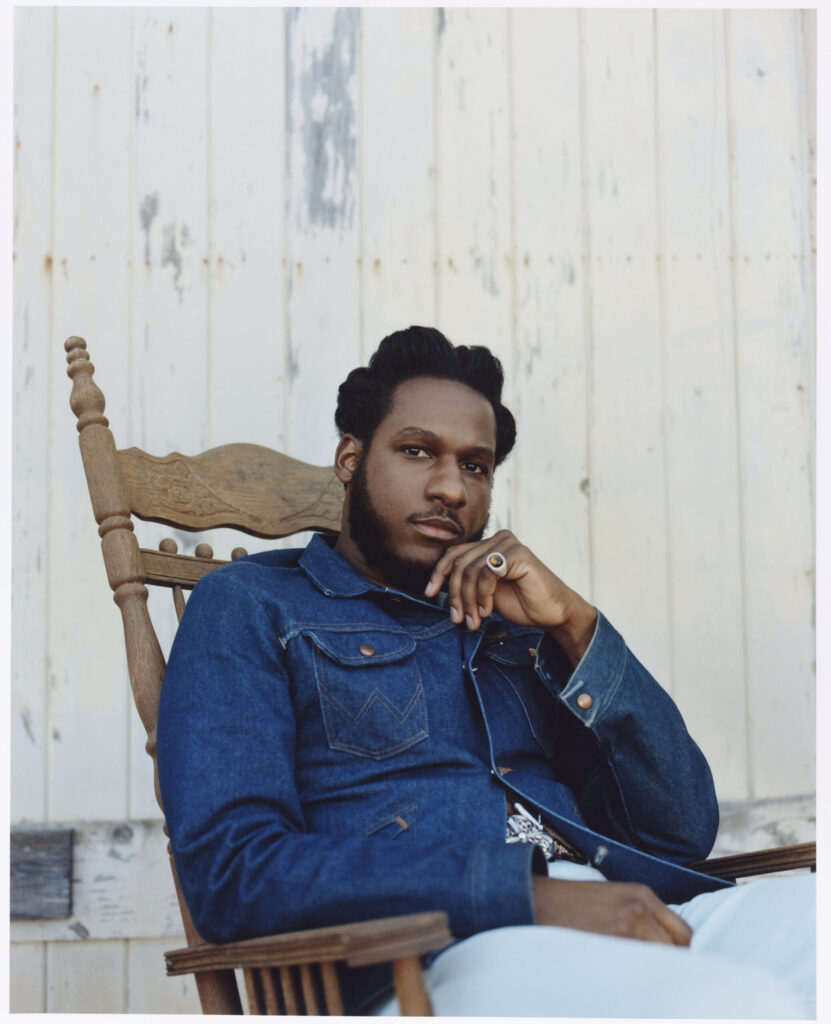 A photo of singer Leon Bridges, wearing a blue denim jacket, sitting in a rocking chair