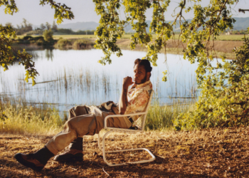 The image features singer Leon Bridges seated in a relaxed pose on a folding chair beside a serene lake, surrounded by grassy fields and trees under clear skies. He is wearing a casual, patterned short-sleeved shirt and light-colored pants, with one hand touching his chin thoughtfully. In the upper left corner, the text "Leon" appears in large letters. The lower right corner contains the Columbia brand logo.