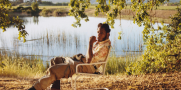 The image features singer Leon Bridges seated in a relaxed pose on a folding chair beside a serene lake, surrounded by grassy fields and trees under clear skies. He is wearing a casual, patterned short-sleeved shirt and light-colored pants, with one hand touching his chin thoughtfully. In the upper left corner, the text "Leon" appears in large letters. The lower right corner contains the Columbia brand logo.