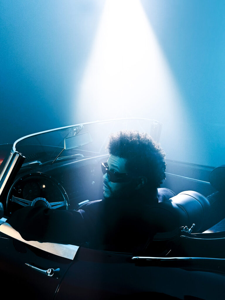 The Weeknd driving a convertible under a spotlight.