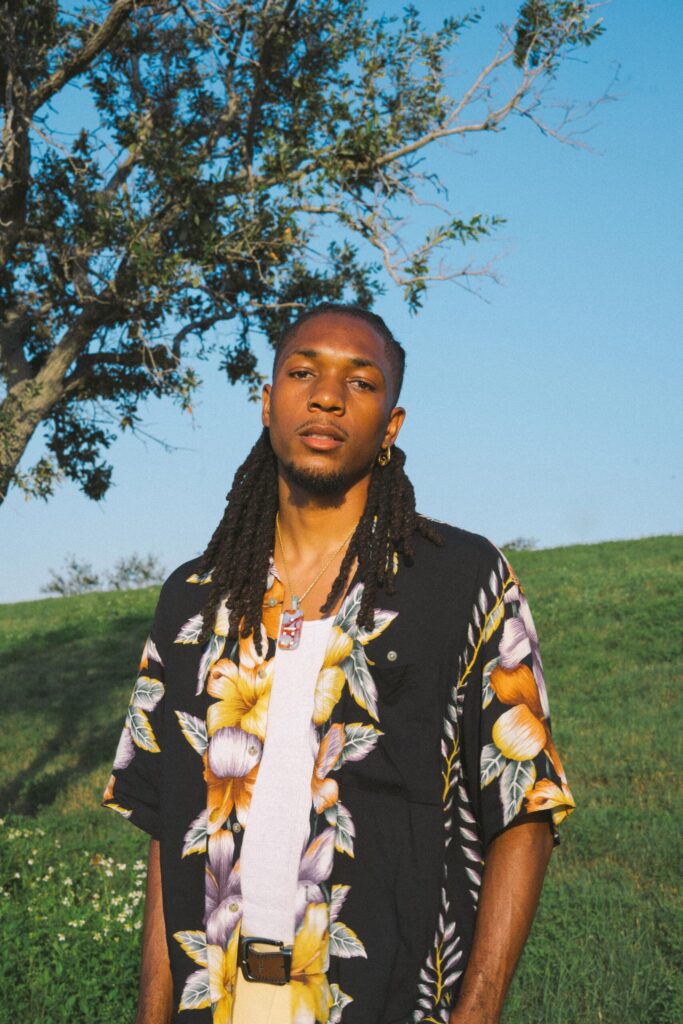 Dominic Scott standing in a field under a tree, wearing a floral shirt and looking directly at the camera with a calm expression.