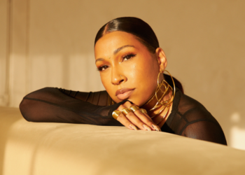 Portrait of a Melanie Fiona resting her head on her arms on a beige couch, lit by warm sunlight. She is wearing a black mesh top and large gold hoop earrings.