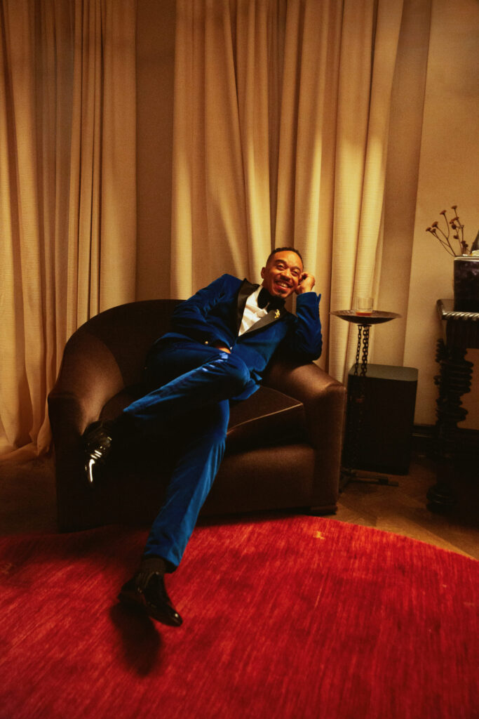 R&B singer lounges in a dark chair, wearing a bright blue suit, in a warmly lit room with elegant curtains and a red rug.