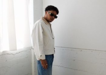 A photo of singer Otis Kane wearing a white jacket and blue jeans, standing in front of a white wall