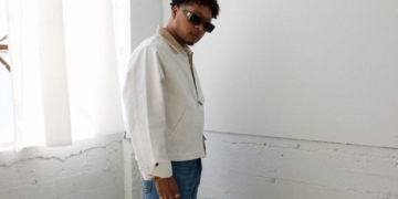 A photo of singer Otis Kane wearing a white jacket and blue jeans, standing in front of a white wall