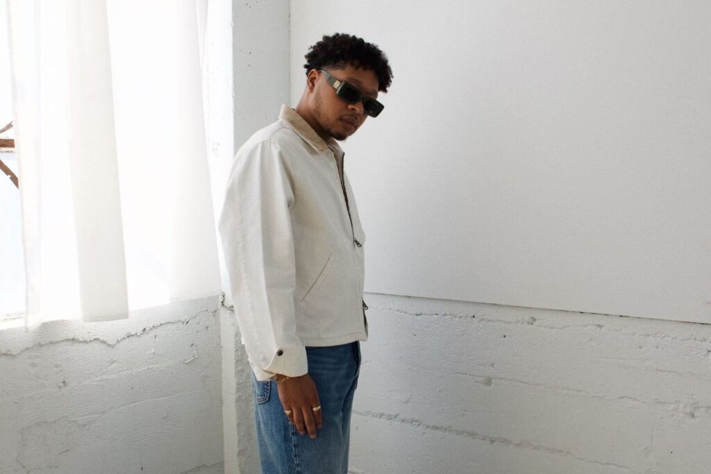 A photo of singer Otis Kane wearing a white jacket and blue jeans, standing in front of a white wall