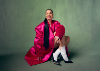 An image of R&B singer Ella Mai in a bright pink trench coat smiling and sitting on the floor against a green backdrop.