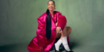 An image of R&B singer Ella Mai in a bright pink trench coat smiling and sitting on the floor against a green backdrop.
