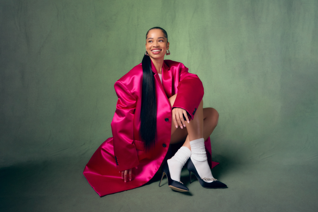 An image of R&B singer Ella Mai in a bright pink trench coat smiling and sitting on the floor against a green backdrop.