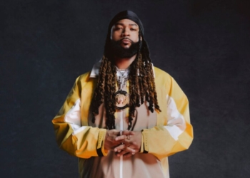 In image of singer PartyNextDoor wearing a yellow, white and tan jacket against a dark grey backdrop.