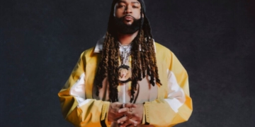 In image of singer PartyNextDoor wearing a yellow, white and tan jacket against a dark grey backdrop.