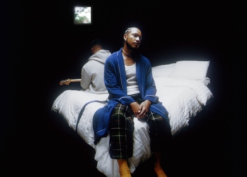 R&B duo Bathe's Inside Voice(s) album cover: Devin Hobdy sits on a bed in a blue robe and pajama pants, while Corey Smith-West, in a gray hoodie, holds a guitar. The bed is dressed with white comforters in a dark room, illuminated solely by light streaming through a small window.