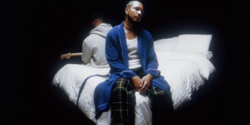 R&B duo Bathe's Inside Voice(s) album cover: Devin Hobdy sits on a bed in a blue robe and pajama pants, while Corey Smith-West, in a gray hoodie, holds a guitar. The bed is dressed with white comforters in a dark room, illuminated solely by light streaming through a small window.