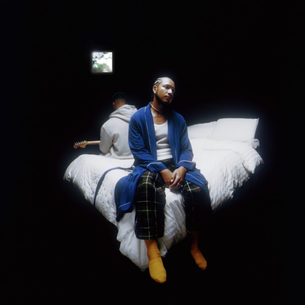 R&B duo Bathe's Inside Voice(s) album cover: Devin Hobdy sits on a bed in a blue robe and pajama pants, while Corey Smith-West, in a gray hoodie, holds a guitar. The bed is dressed with white comforters in a dark room, illuminated solely by light streaming through a small window.
