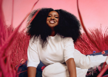 Nao's "Happy People" single cover: Nao with voluminous curly hair, seated on red grass, wearing white and smiling.