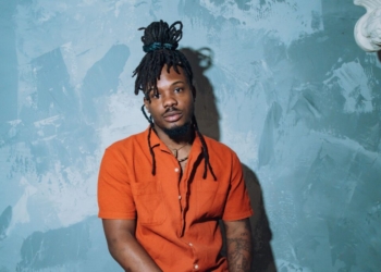 R&B singer Dende sitting in front of a blue wall wearing an orange shirt