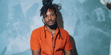 R&B singer Dende sitting in front of a blue wall wearing an orange shirt