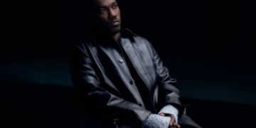 R&B singer Giveon, wearing a leather black jacket, seated in front of a black backdrop.