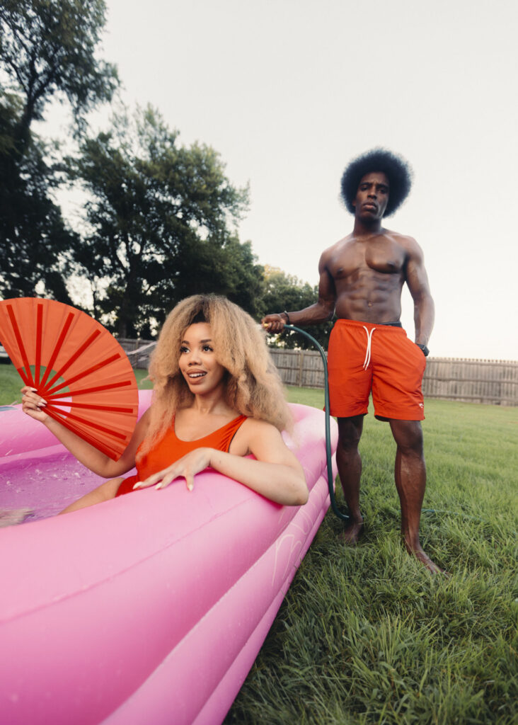 A photo of KIRBY sitting in a pink inflatable pool as Akeem Ali holds a hose filling it up with water